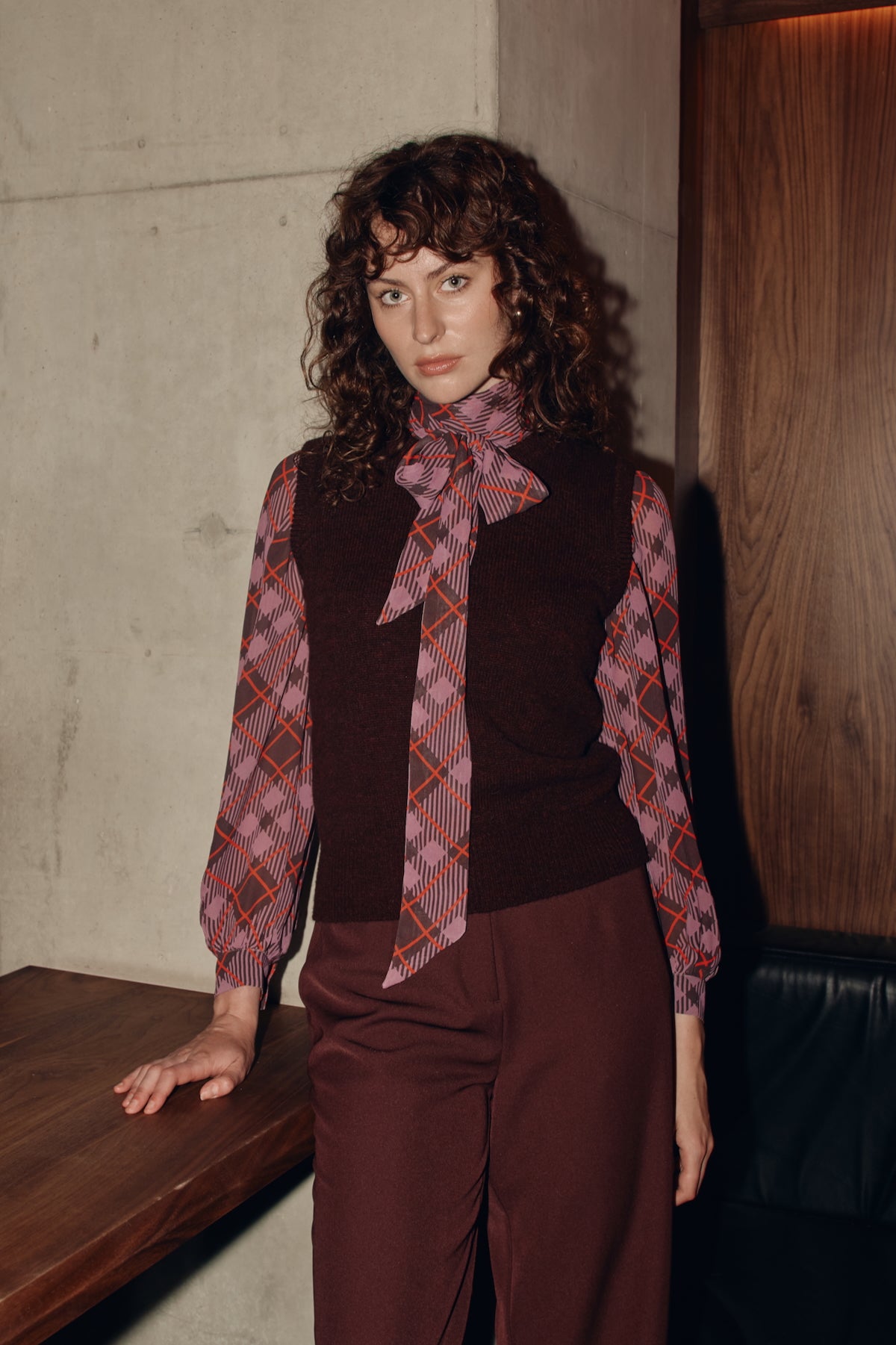 Woman wearing a plaid blouse and burgundy outfit against a textured wall.
