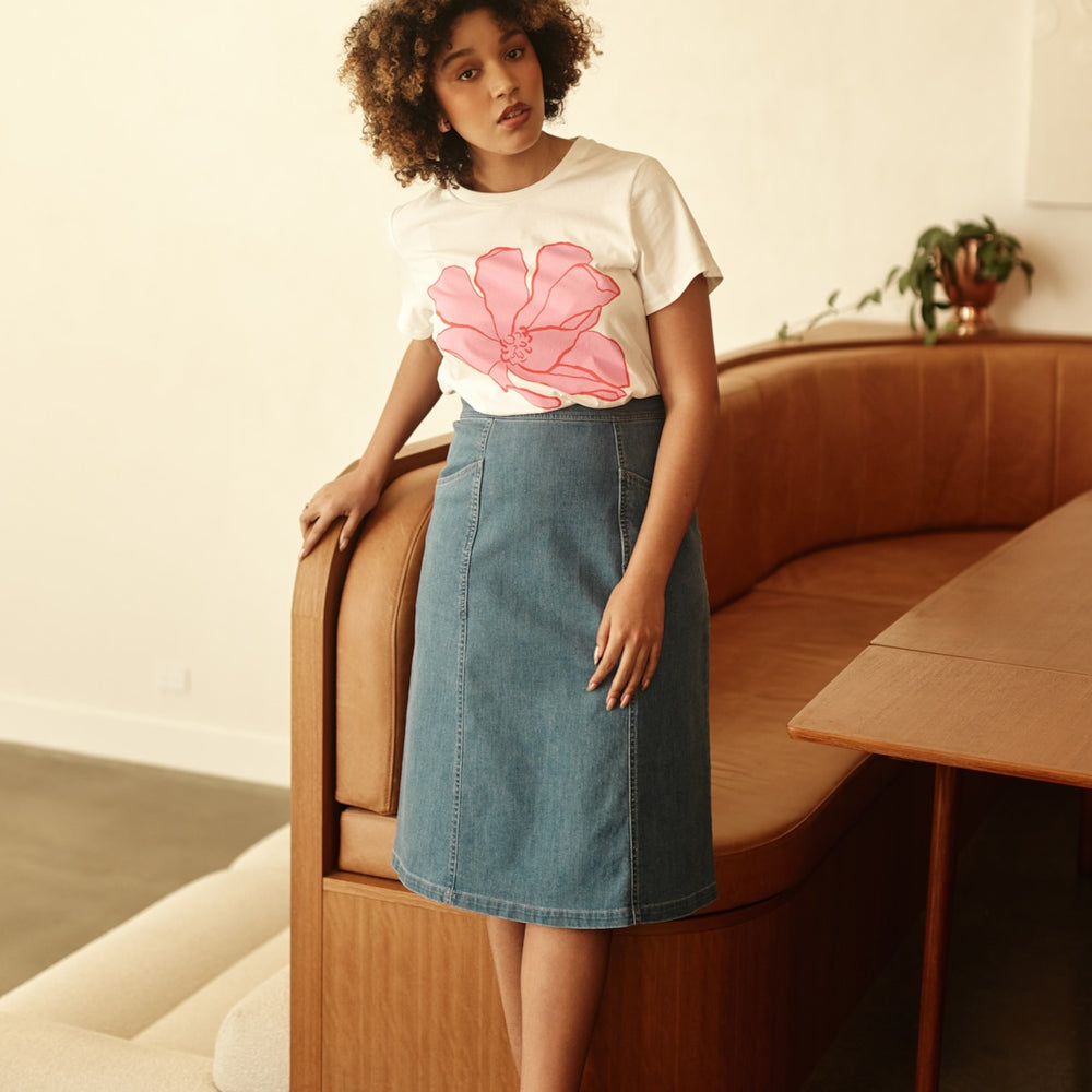 Woman wearing a white t-shirt with a pink flower design and a denim skirt, sitting on a wooden bench.