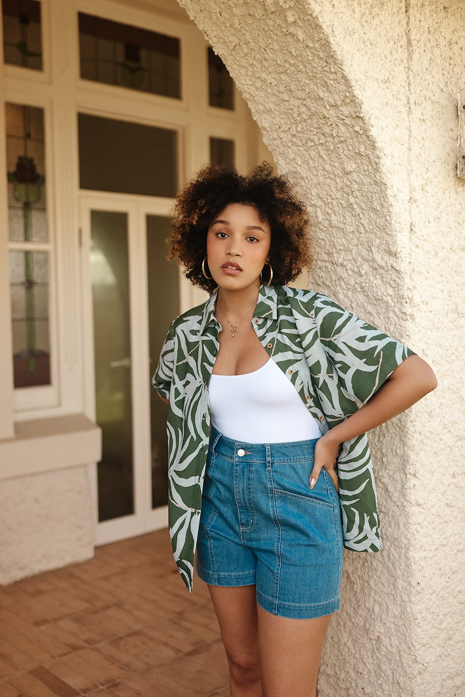 Woman wearing a green leaf-patterned jacket and blue shorts standing against a textured wall.