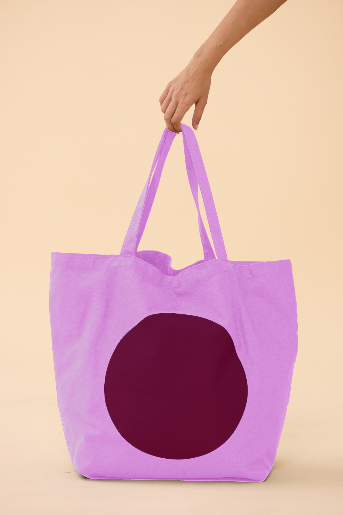 Pink tote bag with a dark maroon circle held by a hand on a beige background