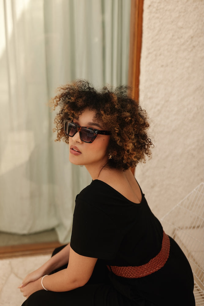 Woman wearing sunglasses and a black dress with a tan belt in a softly lit room.