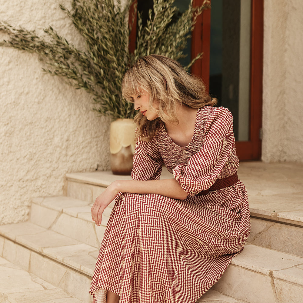 
                      
                        Woman in a burgundy patterned dress sitting on stone steps with a neutral background
                      
                    