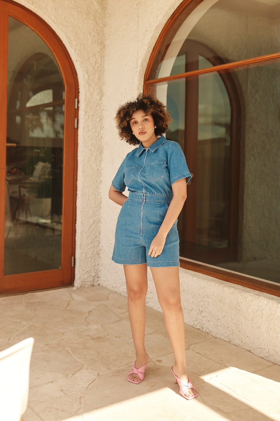 Woman in a short denim jumpsuit standing in front of a building with arched windows.