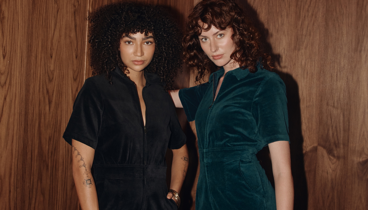 Two women standing against a wooden wall, one in a black velvet jumpsuit and the other in a petrol blue velvet jumpsuit.