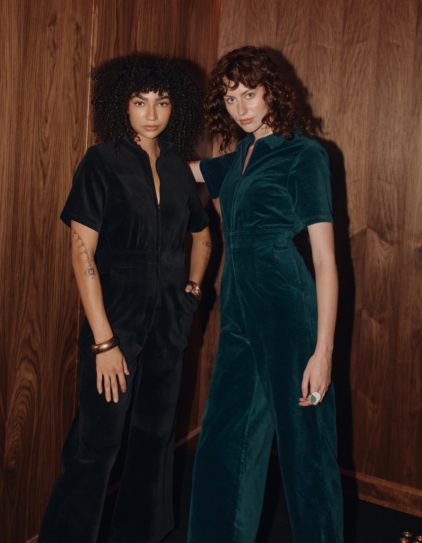 Two women standing side by side against a wooden paneled wall in velvet jumpsuits.