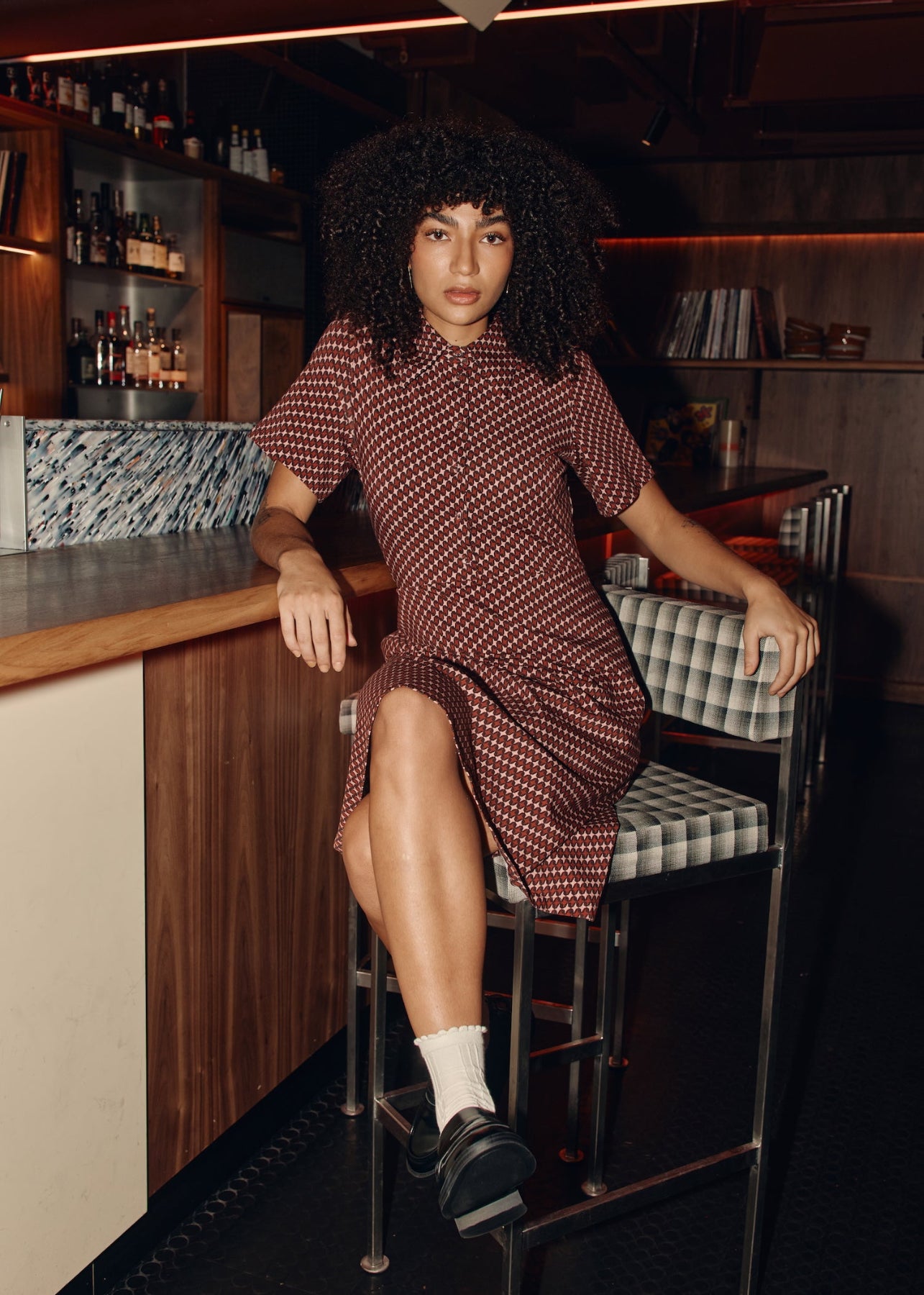 Woman sitting on a bar stool in a bar setting wearing a patterned dress.