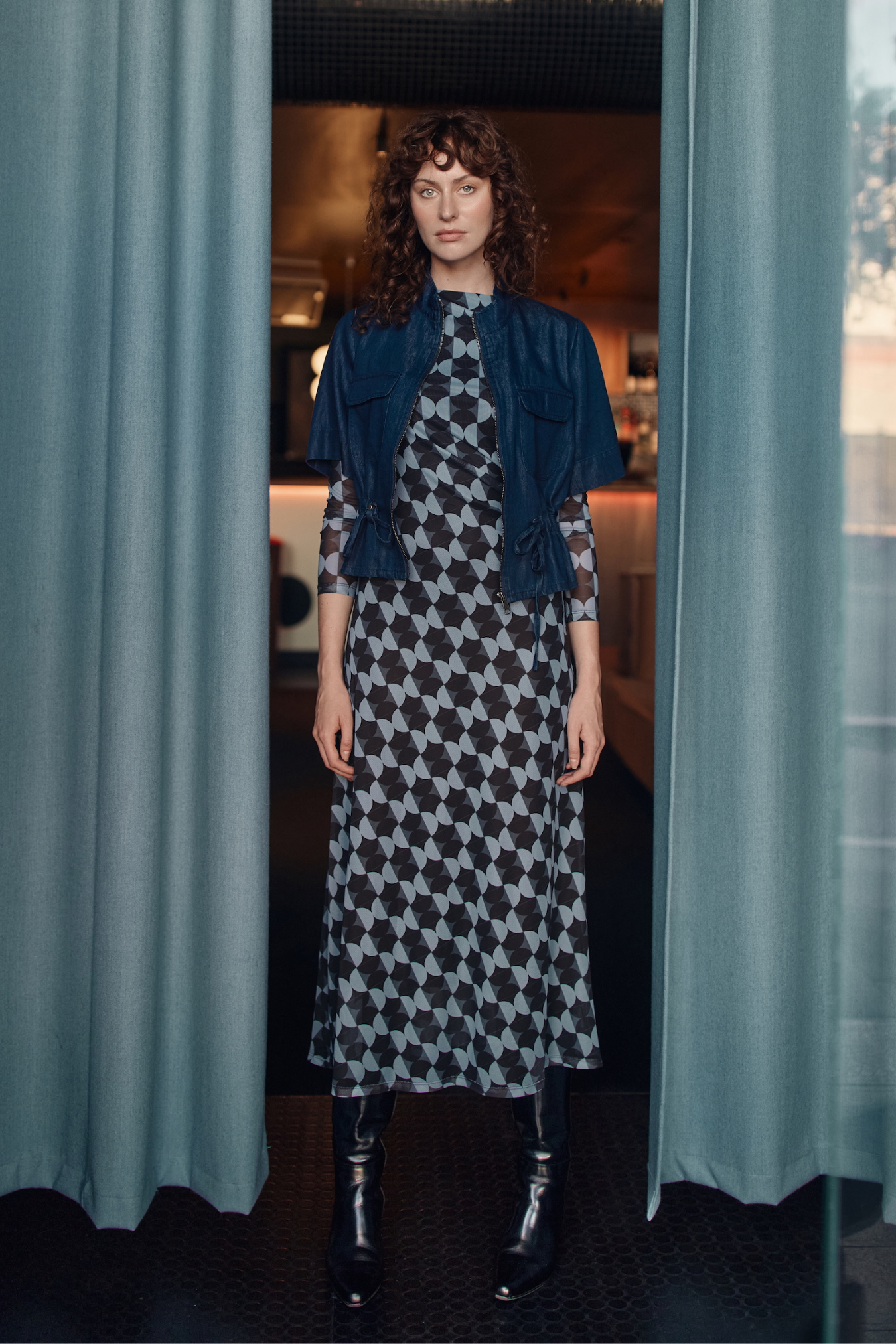 Woman standing between two curtains wearing a patterned dress and chambray jacket.