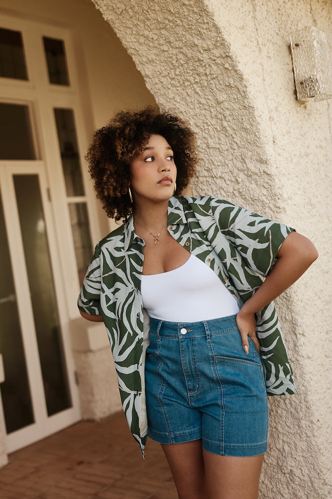 
                      
                        Woman wearing a green and white patterned shirt and blue shorts standing against a textured wall.
                      
                    
