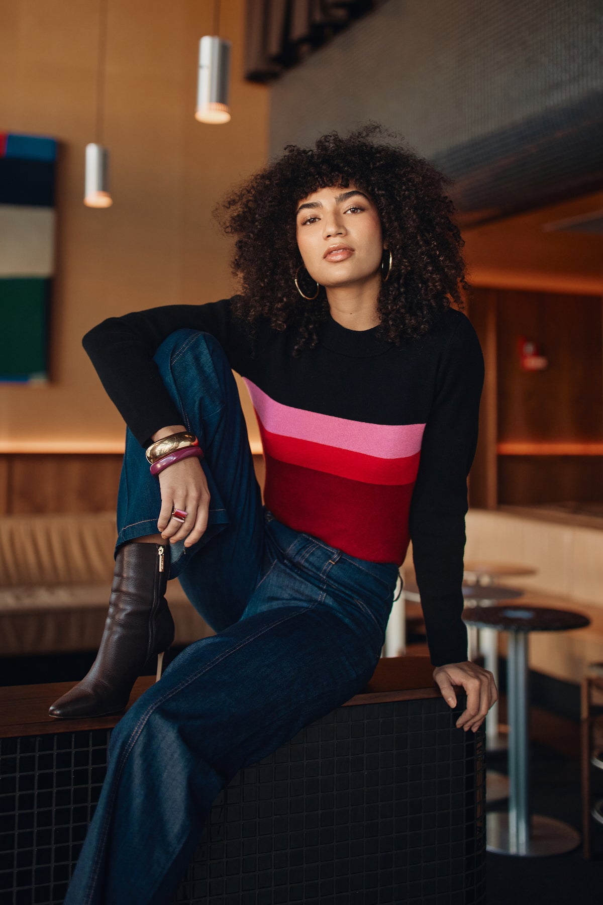 Woman sitting in a modern indoor setting wearing a colorful sweater and blue jeans.