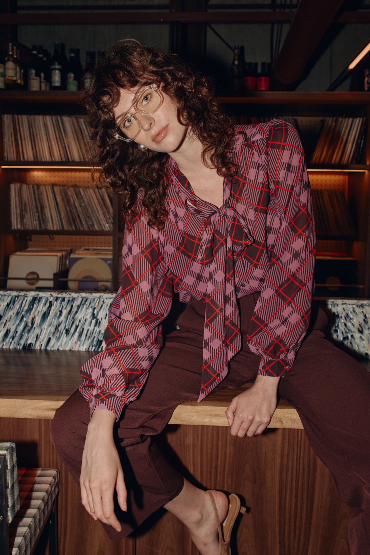 Person wearing a plaid blouse sitting on a record player in a room with vinyl records