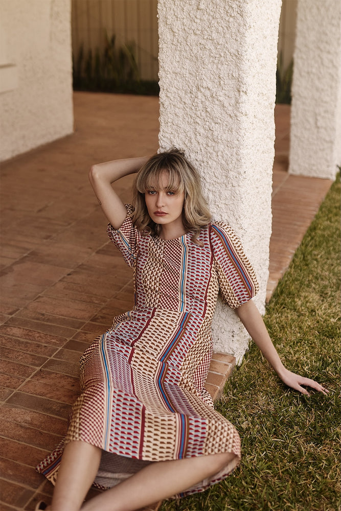 
                      
                        Woman in a patterned dress sitting on grass next to a white column.
                      
                    