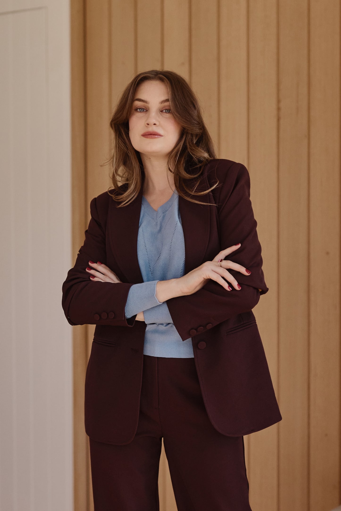 A woman stands with arms crossed wearing a dark burgundy tailored blazer and matching trousers over a light blue blouse, posed against a light wood-paneled wall.