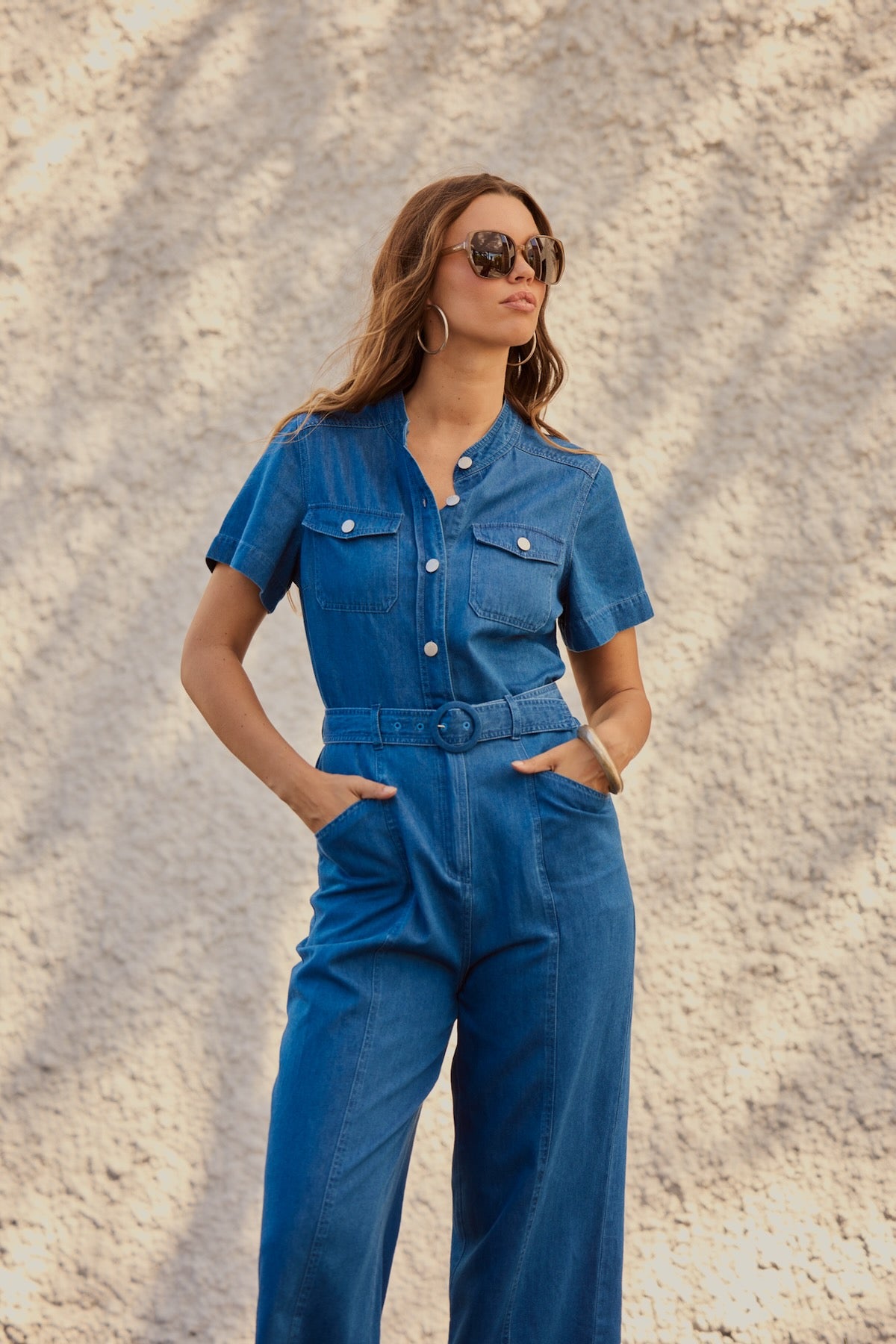 Woman standing in front of a stone wall with oversized glasses on wearing a Frock Me Out CHAMBRAY Safari Jumpsuit
