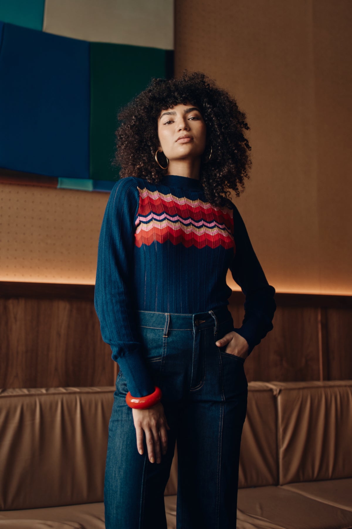 Woman wearing a colorful sweater and jeans standing against a textured wall.
