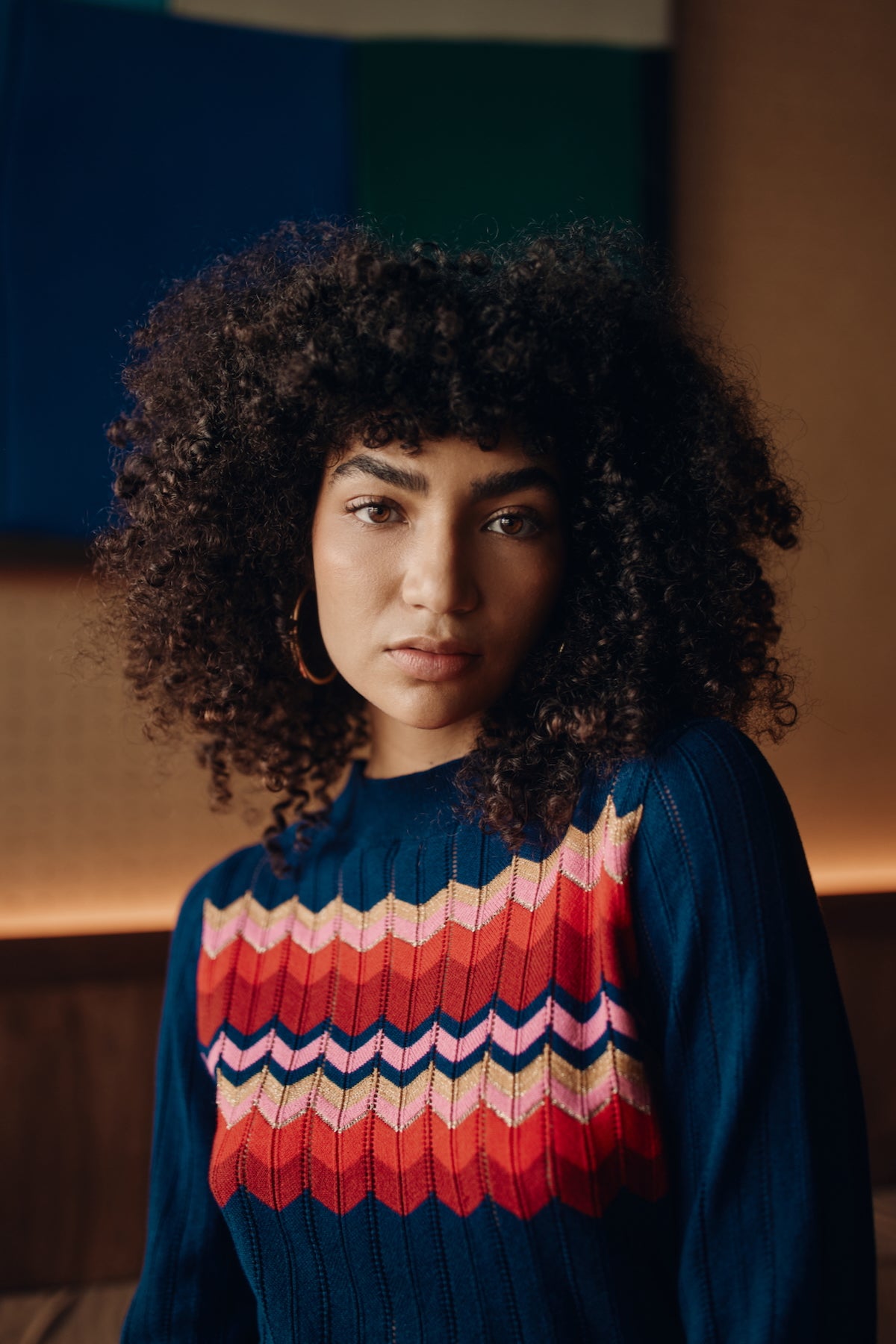 Woman wearing a colorful sweater with a zigzag pattern in a warm-toned room.